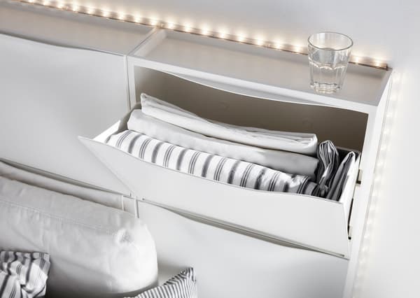 An IKEA TRONES shoe cabinet that’s part of a bed’s headboard is open to reveal fresh and folded bed linens.