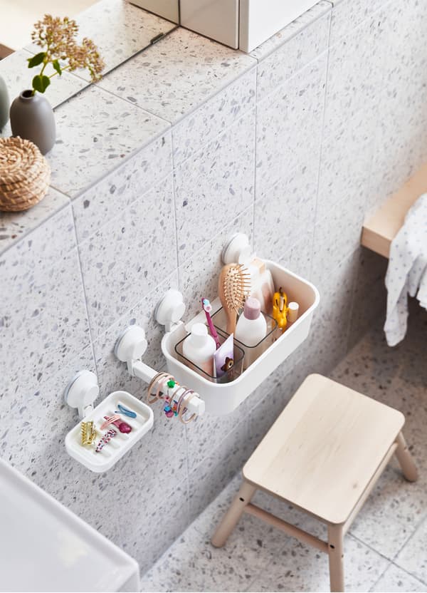 An IKEA VILTO birch step stool and white TISKEN suction cup soap dishes and hooks on a grey and white tiled wall.