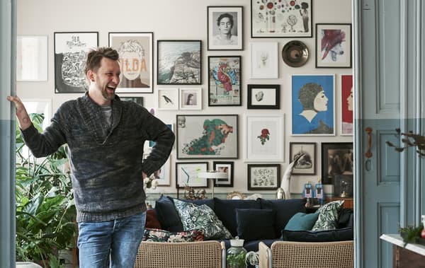 An image of a man standing in his lounge in front of a wall full of pictures.