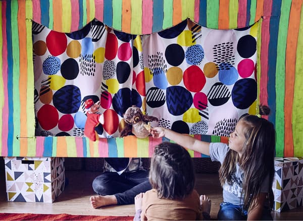An image of two children watching an owl puppet in the puppet theatre and a third child sitting cross legged behind the curtain.