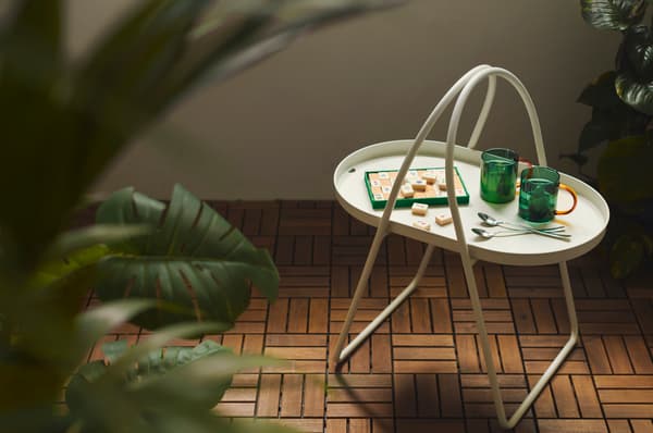 An in/outdoor Monstera FEJKA artificial potted plant outside, standing close to a white MÅBÄRSSKÄR coffee table with two cups, a few spoons and a board game.