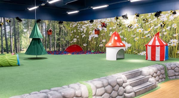 An indoor children’s play area with green flooring, a red-and-white circus tent, and a magical forest backdrop.