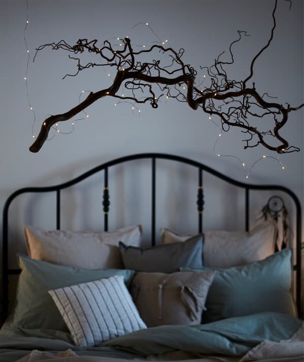 An intricately twisting, leafless, thin tree branch adorned with VISSVASS hung with nylon thread over a cosily made bed.