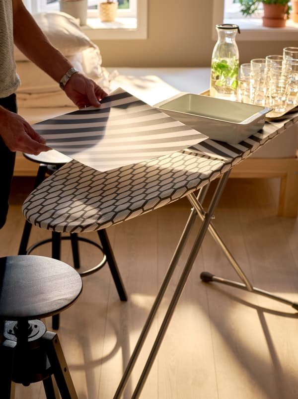 An ironing board being set with serving trays and stacks of glasses and PIPIG place mats protecting the textile surface.