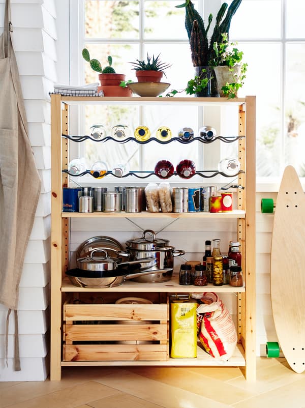An IVAR unit stands by a window, organised as a small pantry with pots, dry goods, canned food, and bottles on racks.