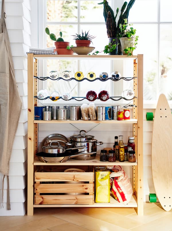 An IVAR unit stands by a window, organised as a small pantry with pots, dry goods, canned food, and bottles on racks.