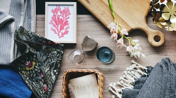 An oak ARTISTISK chopping board, gold-colour STOCKHOLM bowl and some papers in a handmade rattan TRUMMIS basket on a surface.