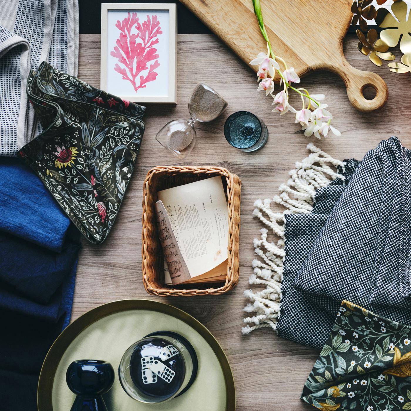 An oak ARTISTISK chopping board, gold-colour STOCKHOLM bowl and some papers in a handmade rattan TRUMMIS basket on a surface.