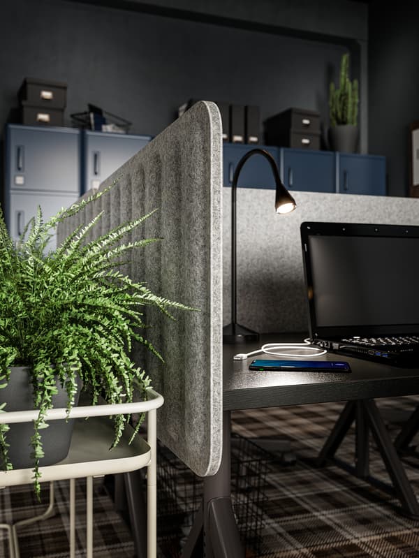 An office interior with EILIF screens in grey attached to a desk with a lit lamp, a laptop and a mobile phone on top.
