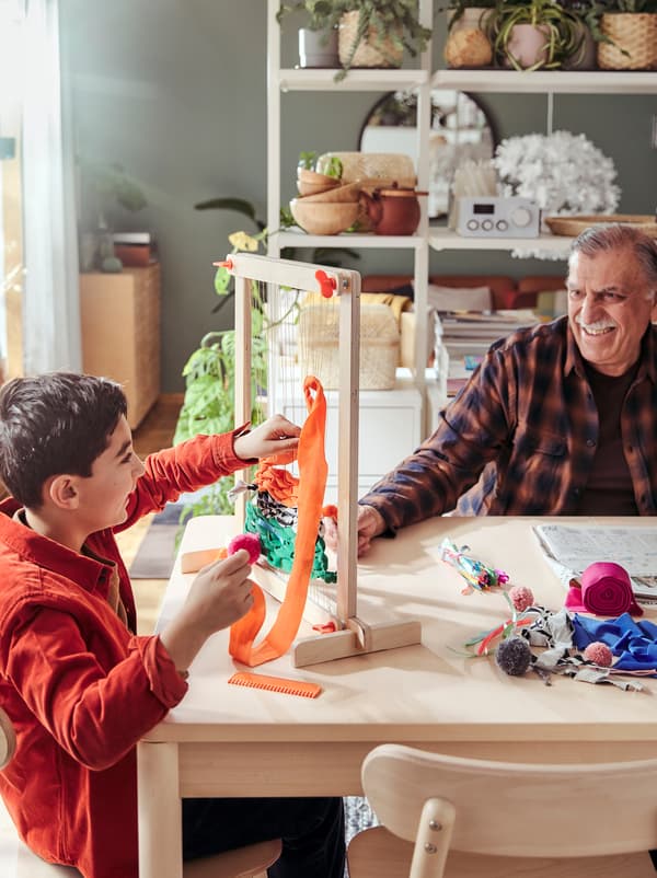 An older man and a young boy smiling at each other from opposite sides of a table on which stands a LUSTIGT weaving loom set.