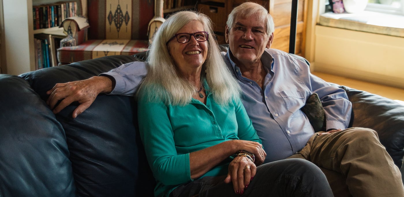 An older woman and man sitting ona  sofa in a living room with a brown patterned chair in the background.