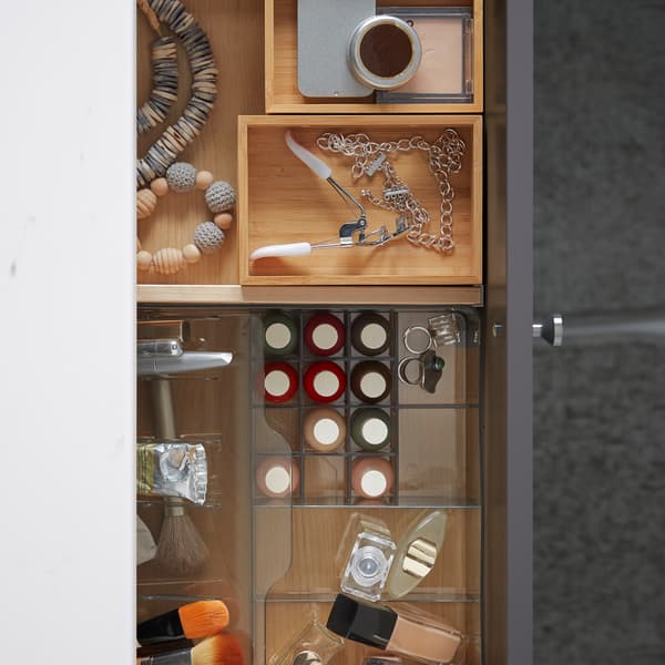 An open bathroom drawer holding an organiser in transparent plastic with make-up, and DRAGAN bamboo boxes with accessories.