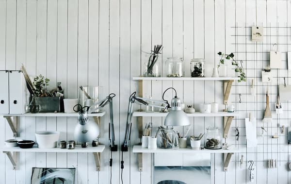 An open cupboard with white shelves holding transparent jars and glassware and a metal shelf holding utensils and papers on hooks.
