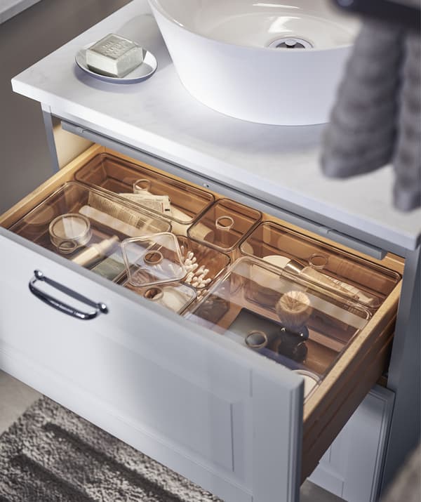An open drawer in a grey washstand unit, inside bathroom items are neatly stored in clear plastic inserts.
