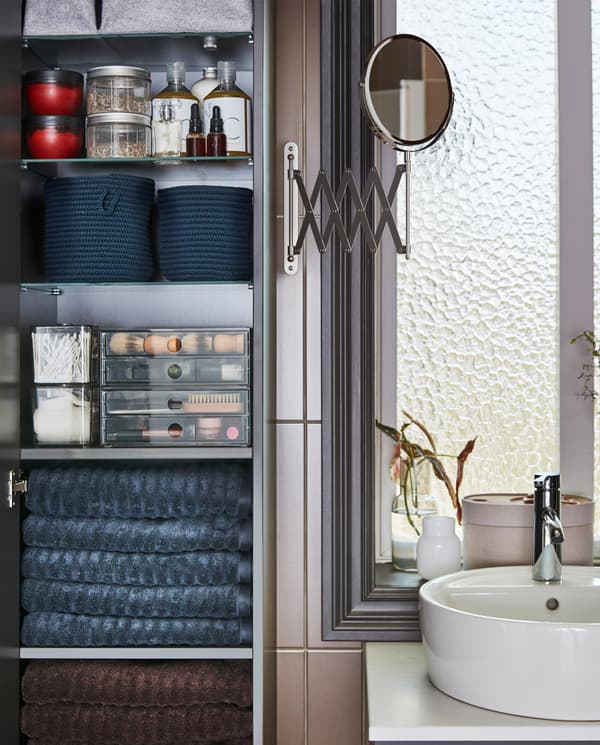 An open high cabinet with dark gray towels, see-through boxes, jars and blue baskets on the shelves and a mirror on the side.