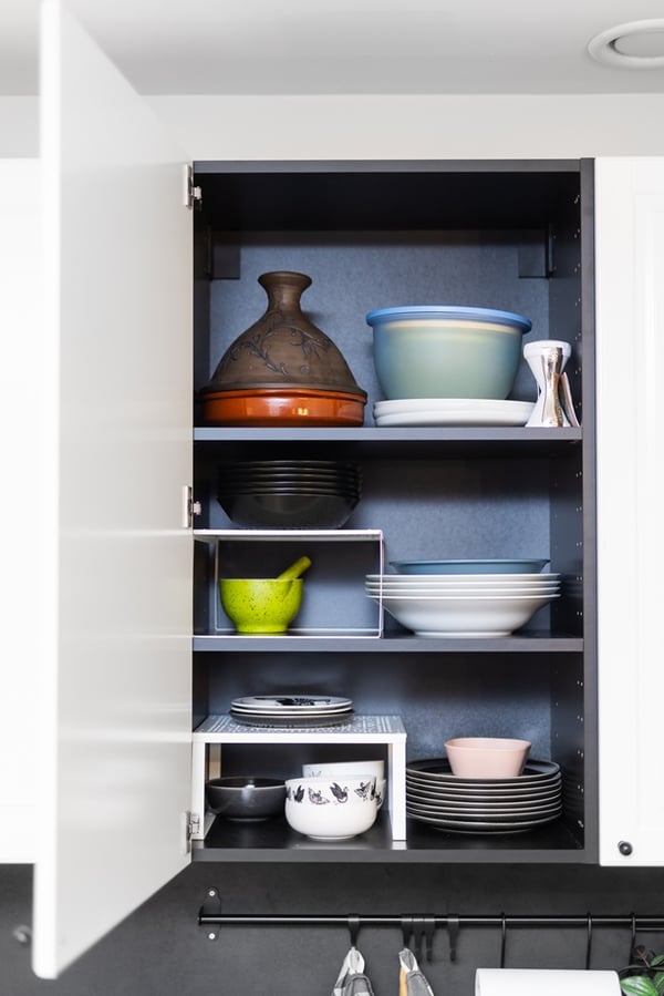 An open kitchen cabinet with shelves holding neatly arranged dishes, bowls, and pots against a dark background.
