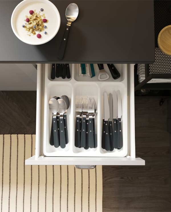 An open kitchen drawer with a cutlery tray with organised cutlery and kitchen utensils, a striped rug and a bowl of yoghurt.