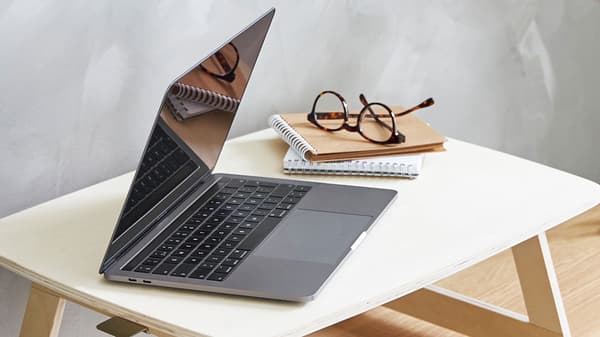 An open laptop on a light-wood laptop table, with a pair of glasses and a notebook next to it. 