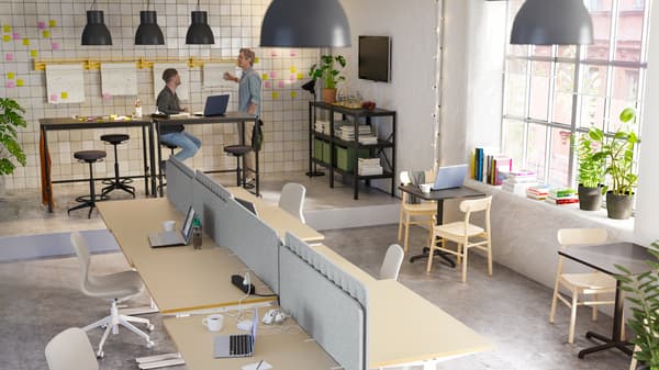 An open office with two men working by high tables, and a woman sitting on a beige/white chair by a desk writing emails.