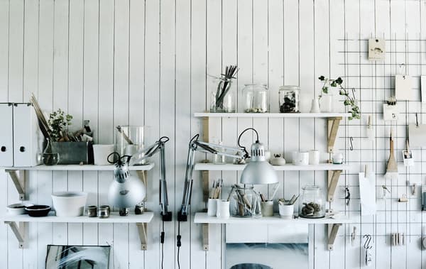 An open pantry with white shelves that hold clear jars and glassware and a wire rack with clip-on utensils and papers.
