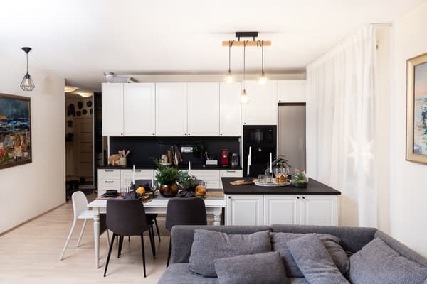 An open-plan kitchen and dining area with white cabinets, a black countertop, a dining table with chairs, and hanging ceiling lights; a gray sofa is visible in the foreground.