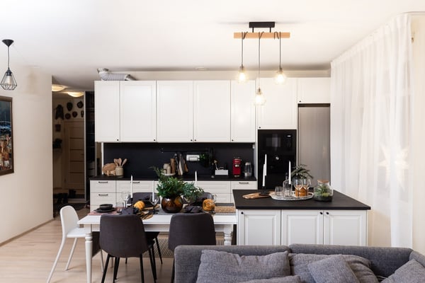An open-plan kitchen and dining area with white cabinets, a black countertop, a dining table with chairs, and hanging ceiling lights; a gray sofa is visible in the foreground.