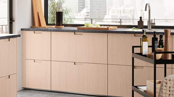 An open-plan kitchen with FRÖJERED fronts in light bamboo. There is a NISSAFORS trolley with condiments and boxes.