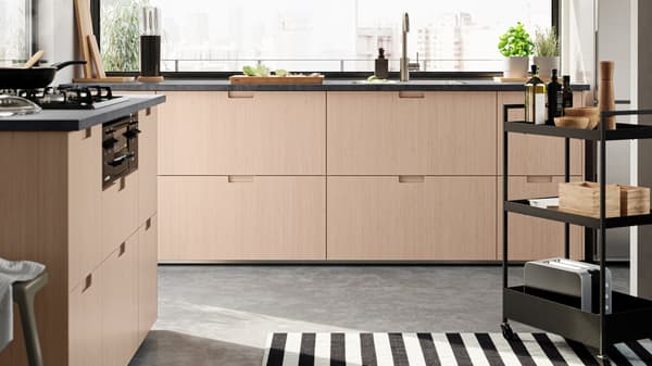 An open-plan kitchen with FRÖJERED fronts in light bamboo. There is a NISSAFORS trolley with condiments, boxes and a toaster.