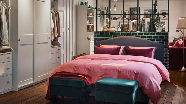 An open-plan space with a HAUGA bed covered in pink bed linen and with two green footstools at the end.