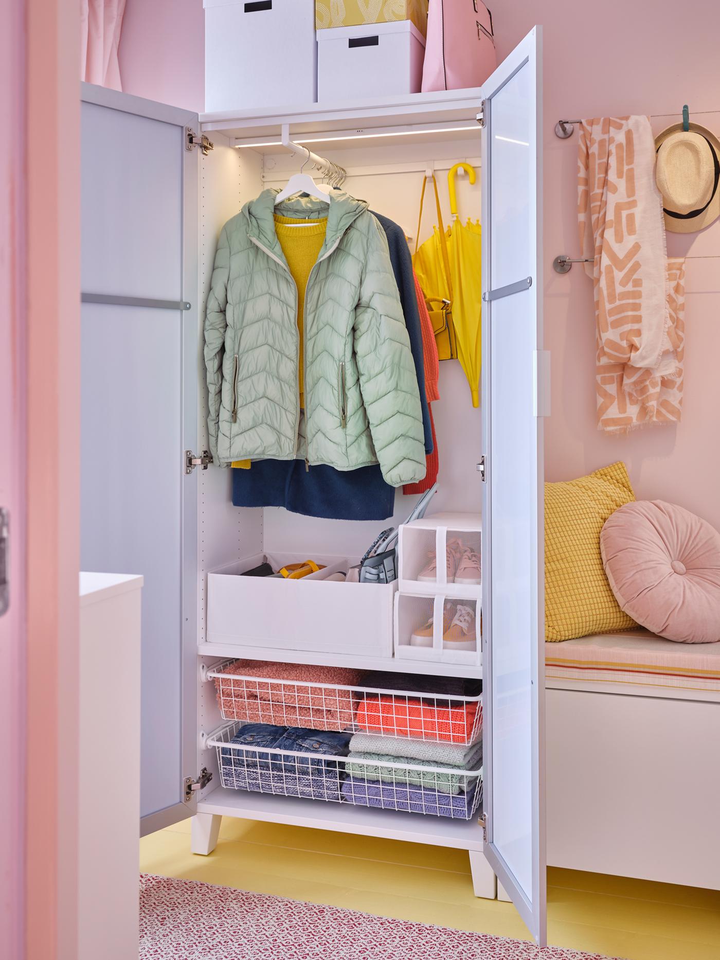 An open PLATSA wardrobe in a pink hallway showing baskets, shelves, hooks and a hanging rail with a green coat on it.