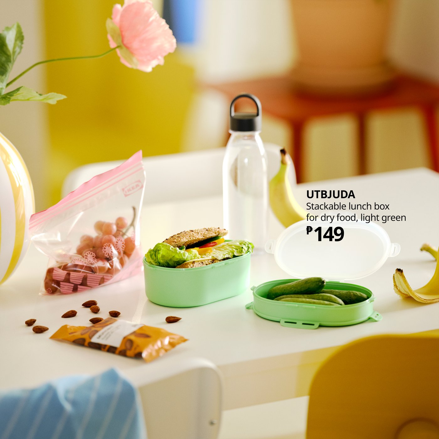 An open UTBJUDA stackable lunch box contains a sandwich in one section and pickles in the other. A water bottle is behind.