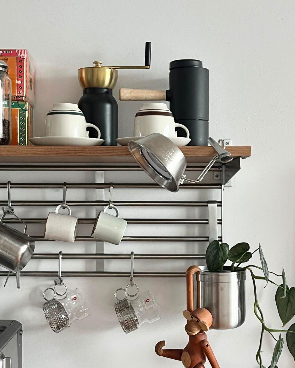 An open wall shelf above a coffee corner, with hanging coffee cups, a manual coffee grinder and a thermos on the shelf, metal mugs on hooks, a small hanging plant pot, and a wooden decorative figure against a light-coloured wall.
Geef uw feedback over BizChat