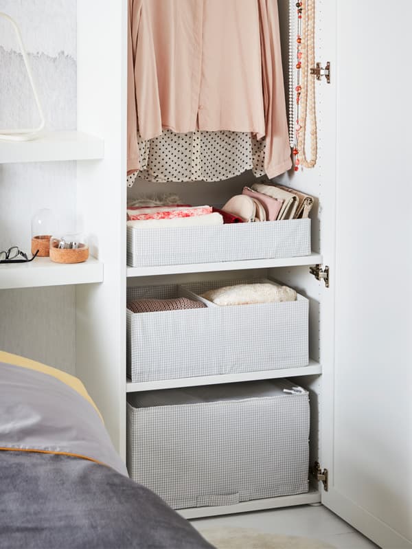 An opened white IKEA PLATSA wardrobe, next to a bed, revealing clothing and white patterned storage boxes.