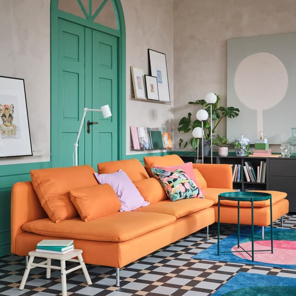 An orange 4-seat sofa with chaise longue, a green tray table, two rugs in pink/green/blue and two floor lamps in white.