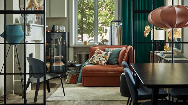 An orange sofa bed in the middle of a living room with gray walls, green curtains, a glass door cabinet and a beige rug.