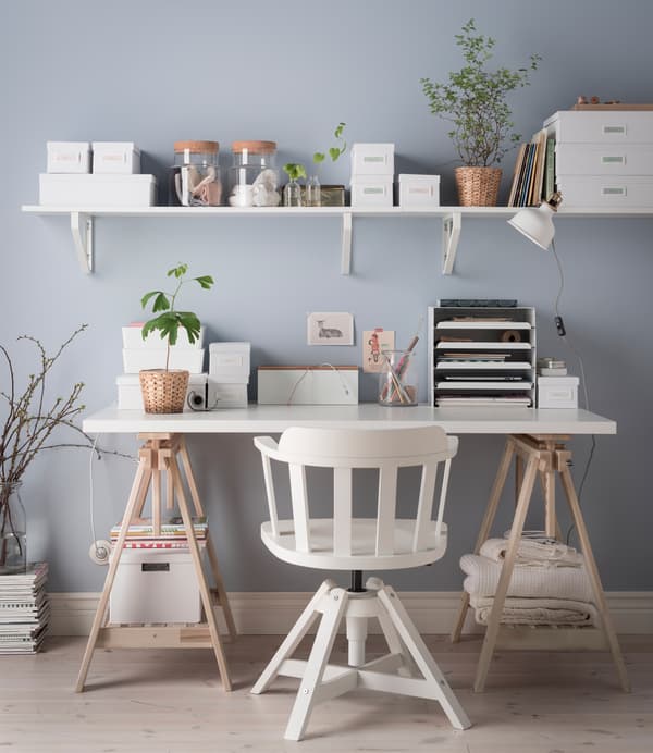 An organized desk without drawers in white and light wood with a shelf above it and lots of boxes, jars and vases for storage.