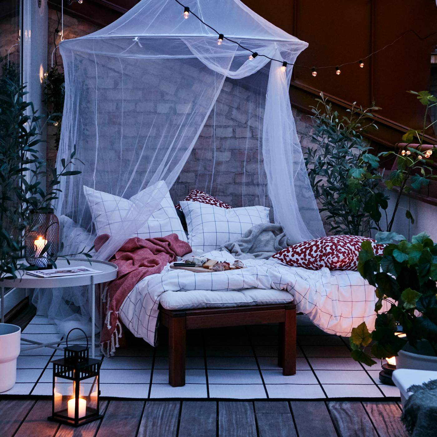 An outdoor area with a brown stained sun lounger made as a bed with white/black checked bed linen on a rug under a white net.