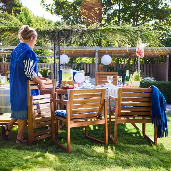 An outdoor dining area features a light brown stained NÄMMARÖ chair with armrests around a wooden table set for a meal.