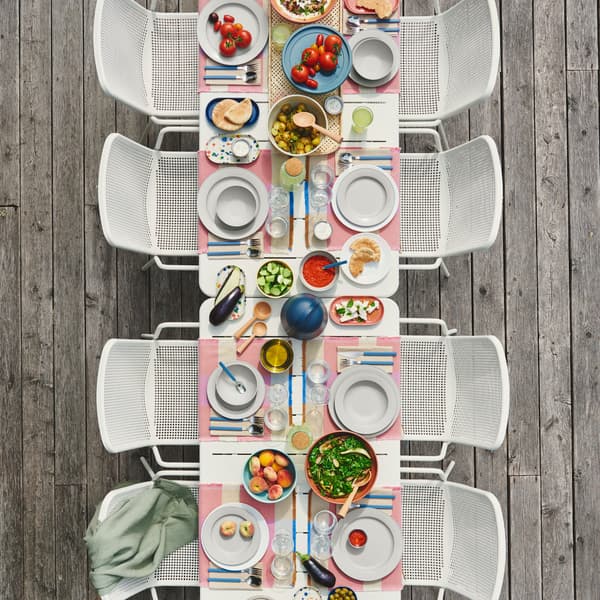 An outdoor dining table for eight is set with green-grey SIMKRABBA plates, and pastel dishes and glasses arranged around.