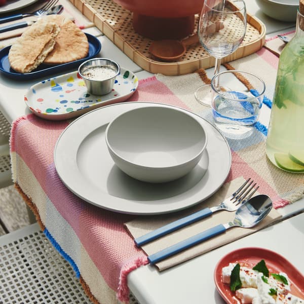 An outdoor dining table is set with a green-grey SIMKRABBA plate, with various dishes and glasses arranged around.