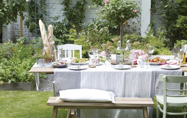 An outdoor dining table set for lunch with a grey and white table cloth and white tableware. 