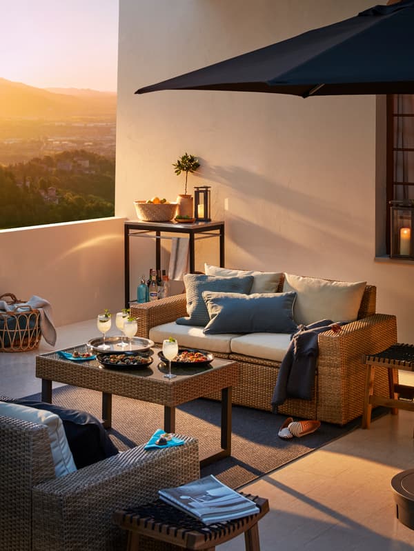 An outdoor sofa with white cushions on a terrace with a parasol, coffee table with refreshments and an outdoor armchair.