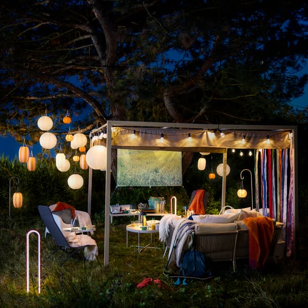 An outdoor space at night featuring striped solar-powered orange and white SOLVINDEN pendant lamps hanging from a large tree.