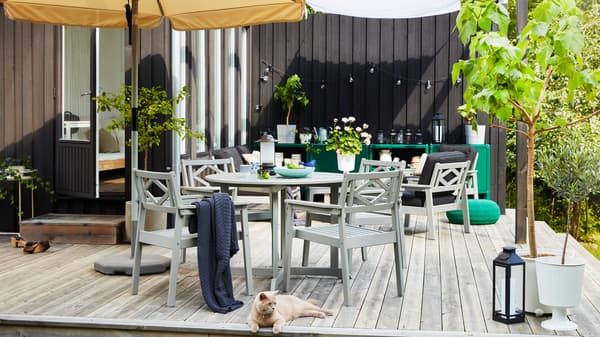 An outdoor space on a wooden deck with a beige parasol, grey outdoor furniture, green planted trees and a cat.
