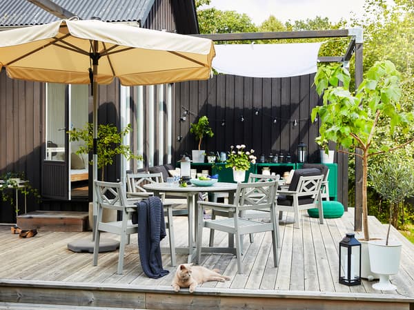 An outdoor space on a wooden deck with a beige parasol, grey outdoor furniture, green planted trees and a grey/orange cat.