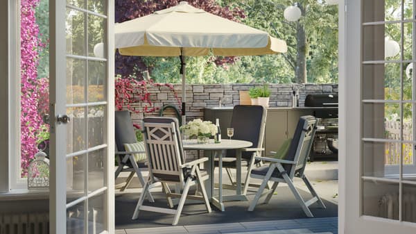 An outdoor table with a beige parasol, reclining chairs and dark grey cushions, brick wall fence and outdoor kitchen.