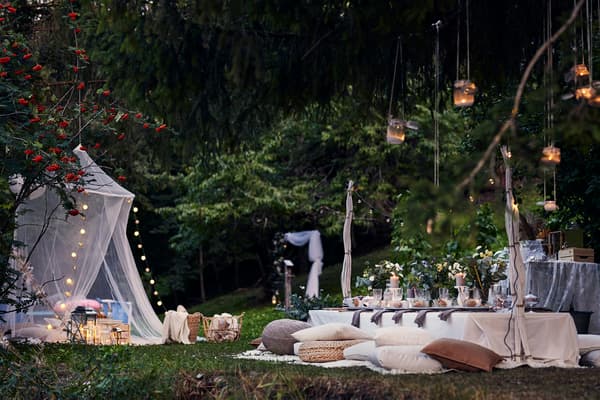 An outdoor wedding set up with fairy lights hanging from a white canopy, a low picnic table with cushions around it and lanterns hanging from the trees.