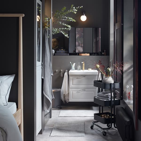 An overall grey bathroom with a wash stand and high cabinet in light grey, a black trolley and many dried decorative flowers.