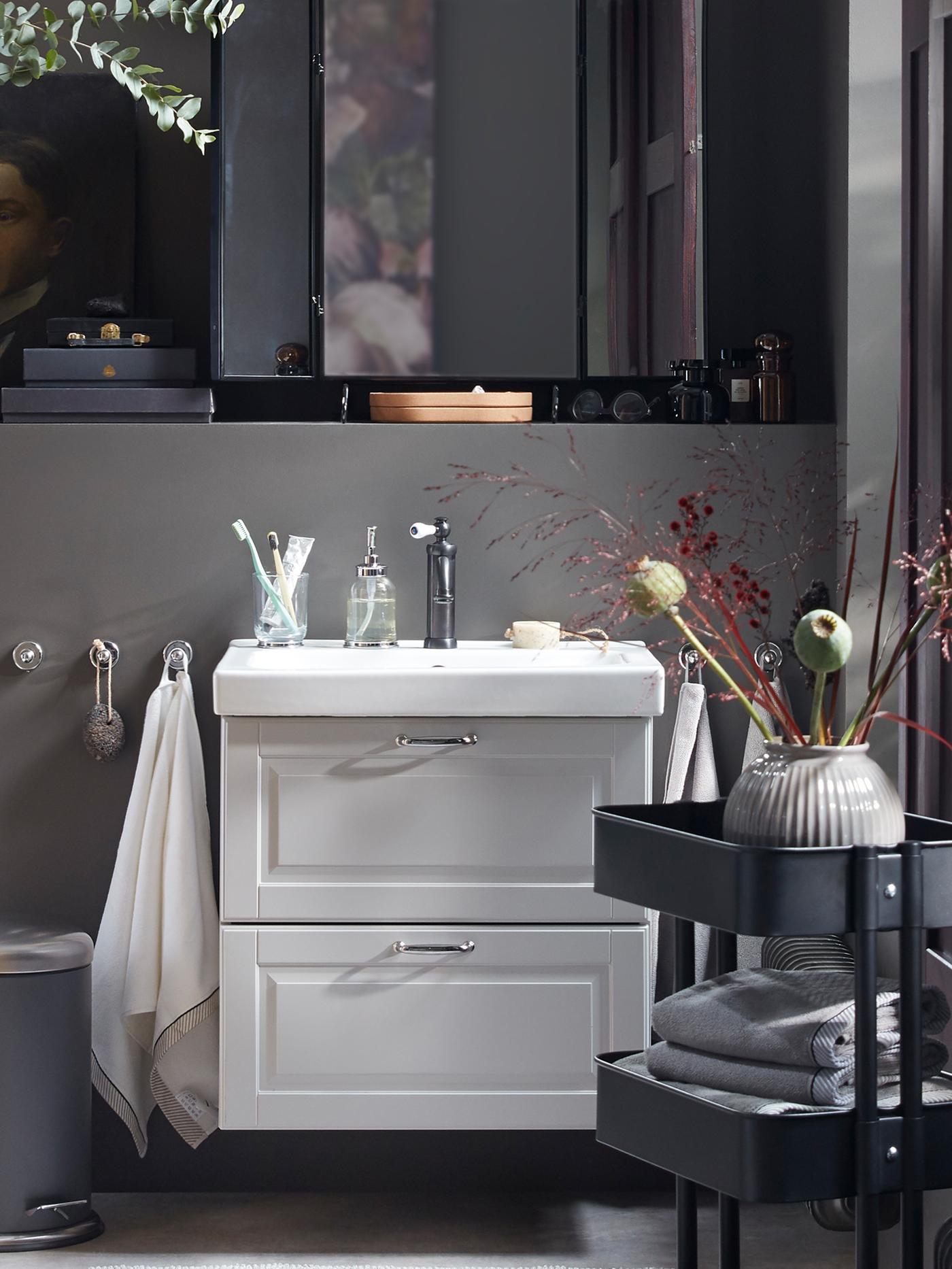 An overall grey bathroom with a wash-stand with bathroom articles on it, a black trolley and many dried decorative flowers.