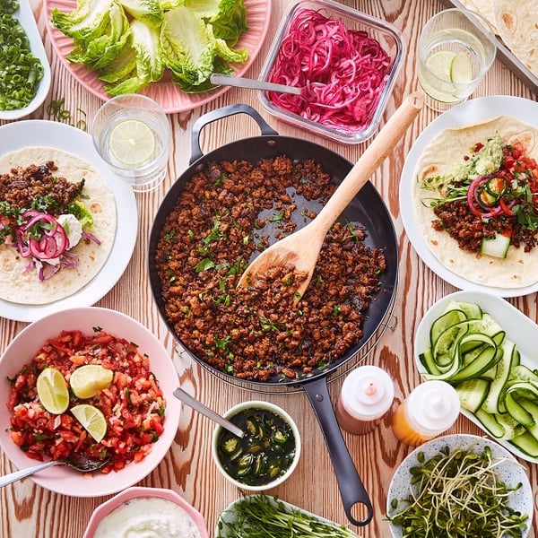 An overhead view of a colourful table spread featuring a central pan of cooked plant-rich mince surrounded by bowls of fresh vegetables, sauces and toppings.